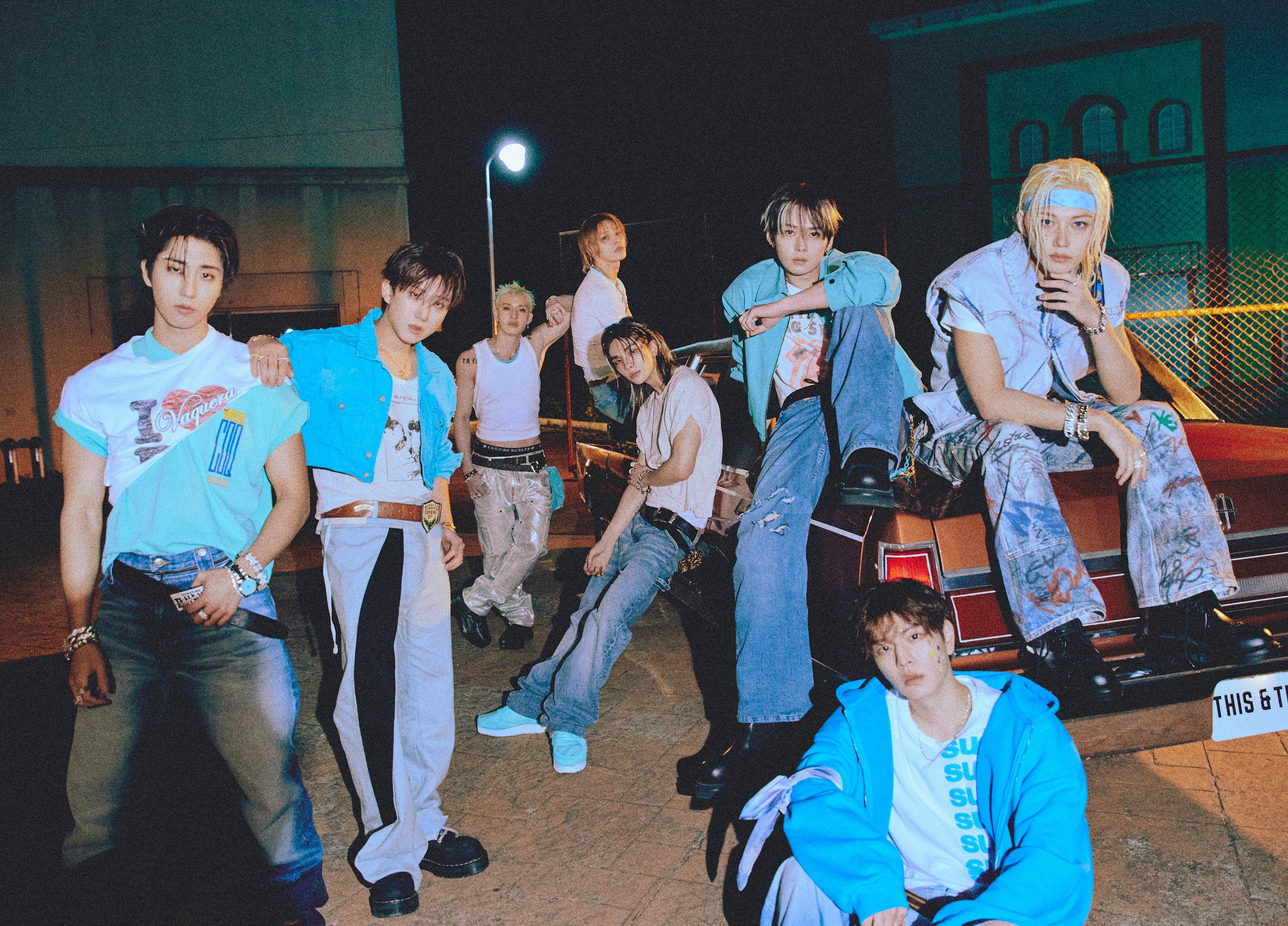 A group of seven young men posing together in a nighttime urban setting, wearing various colorful streetwear outfits including blue, white, and gray tones.